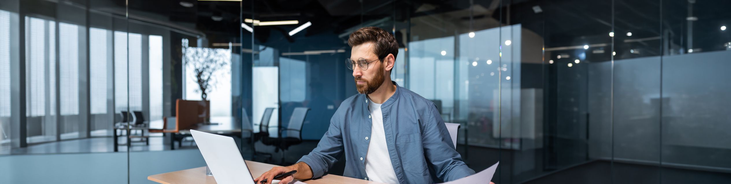 Mann arbeitet in einem modernen Büro an einem Laptop, hält ein Dokument in der Hand und blickt konzentriert auf den Bildschirm.