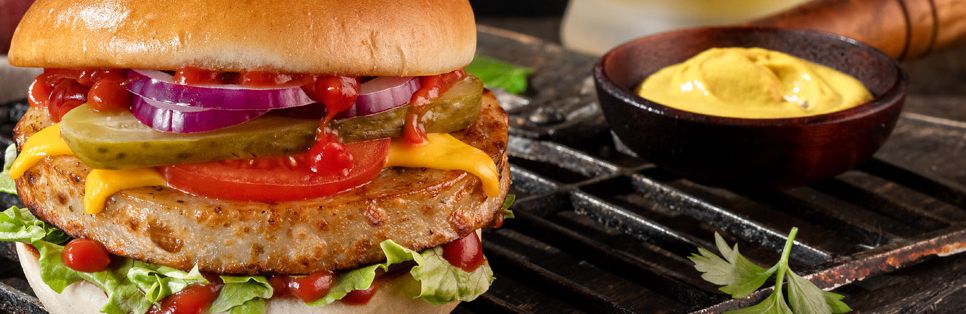 Belegter Burger mit Bratwurst-Patty, Zwiebeln, Tomaten, saure Gurken, Salat und Ketchup liegt auf einem Rost, daneben steht ein kleines Schälchen mit Dip.